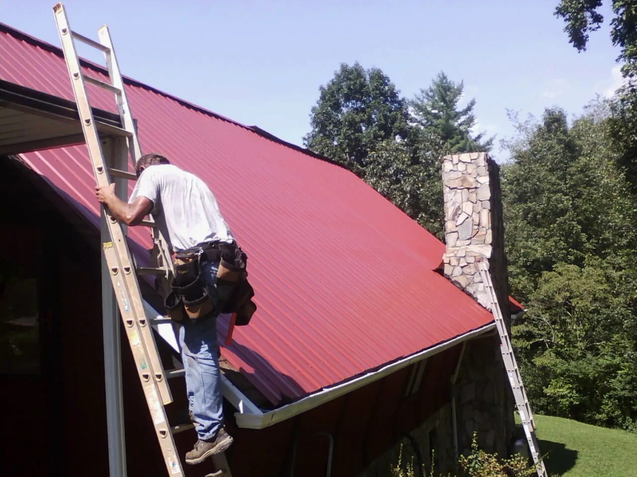 Roofer on a ladder beside a red metal roof and stone chimney — Soffit & Fascia Repair by Roofing Brothers in Berry